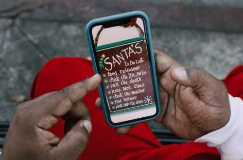 Close-up of hands holding a smartphone displaying Santa's whimsical to-do list outdoors.